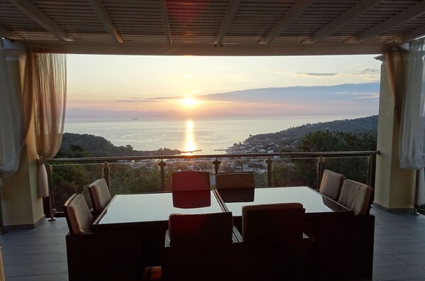 A dining area with a table and chairs, overlooking a scenic view of the sea and hills during sunset.