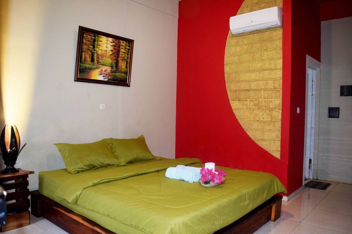 A hotel room with a bed, green bedding, a painting of a forest, a red accent wall, and an air conditioning unit.