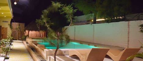 Un área de piscina nocturna con sillones de descanso y un pequeño árbol.