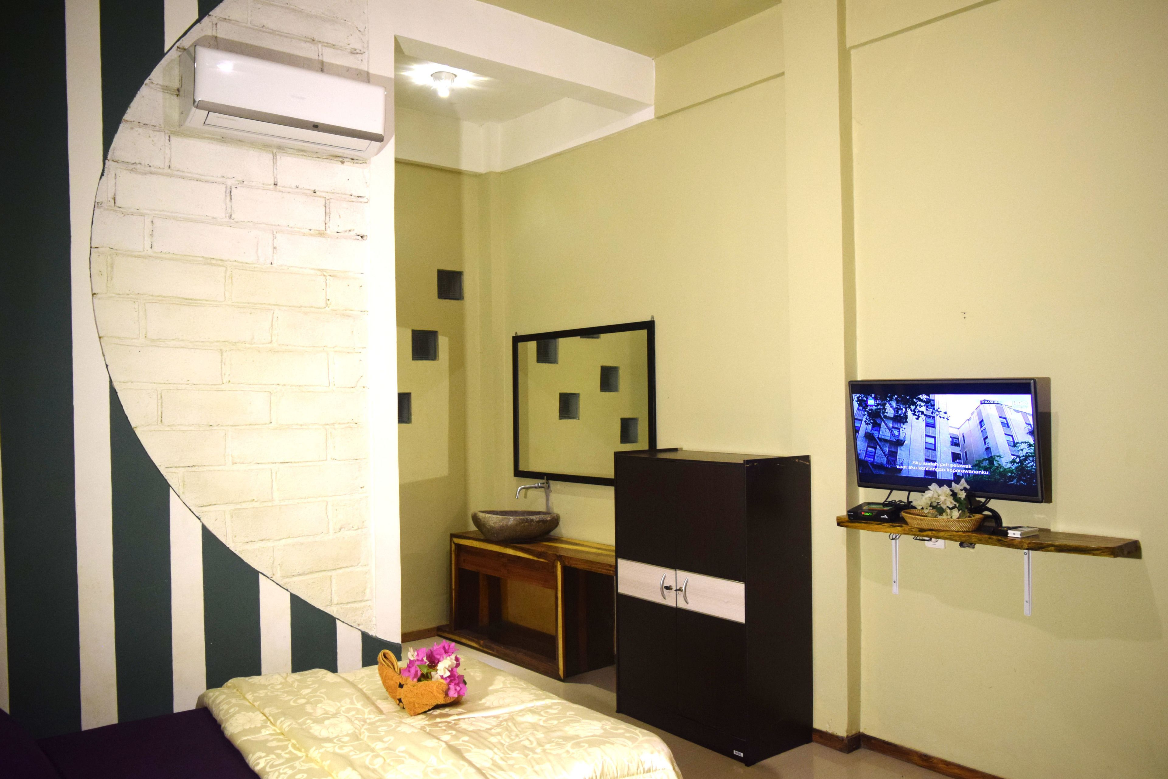 Una habitación de hotel con una pared de ladrillo curva, una cama con almohada floral, una consola de madera con lavamanos en cuenco, un mueble negro y un televisor montado en la pared.