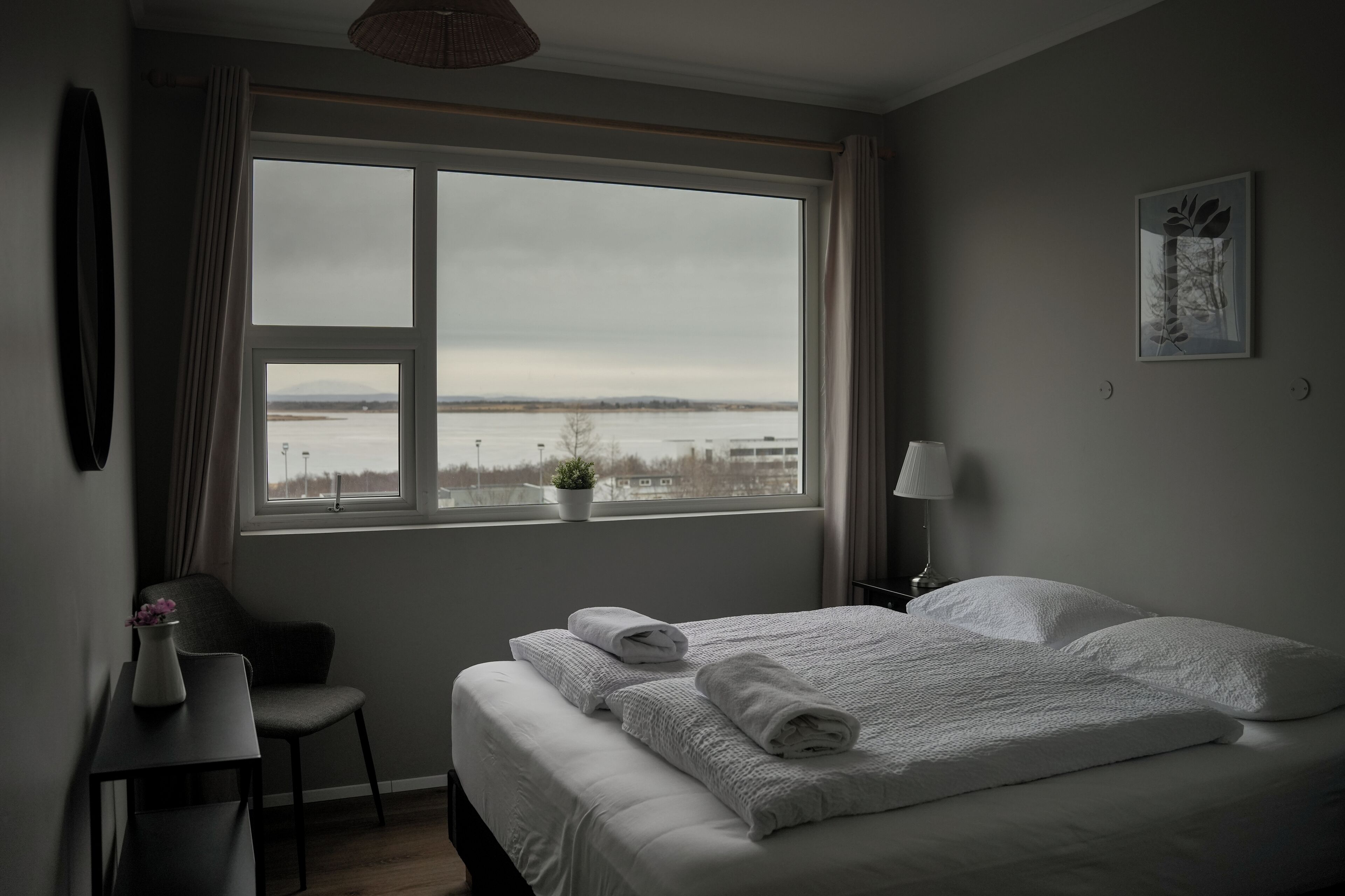 A bedroom with a large window, a bed with white linens, a chair, a small table, and a wall-mounted lamp.