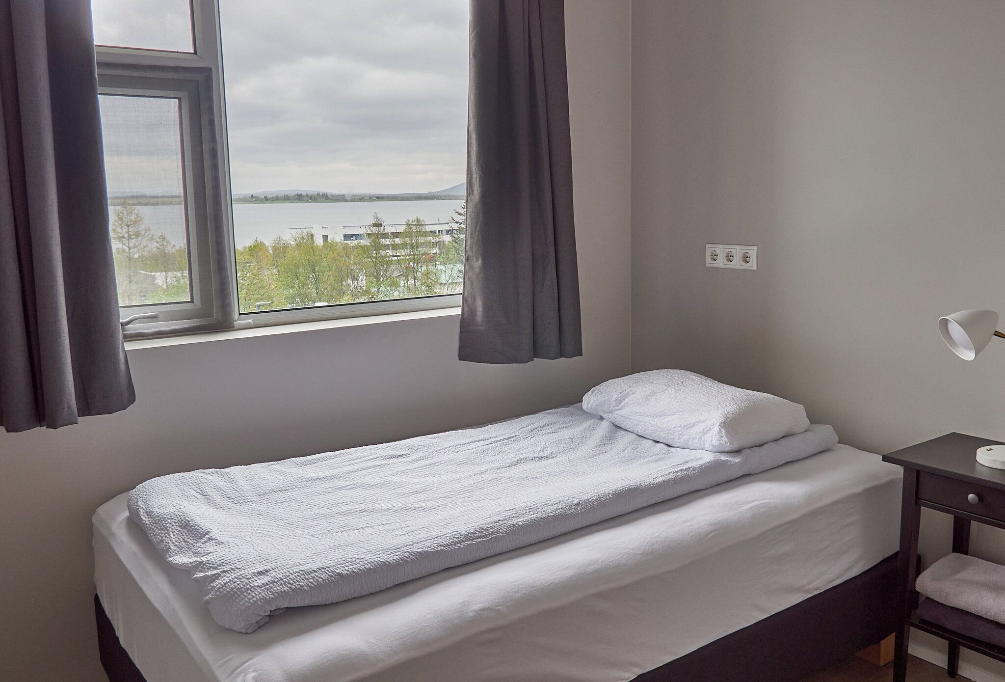 Una cama individual con ropa blanca, una ventana con cortinas, una mesita de noche con lámpara y vistas a un cuerpo de agua y árboles.