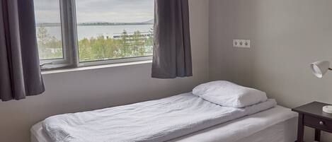 Una cama individual con ropa blanca, una ventana con cortinas, una mesita de noche con lámpara y vistas a un cuerpo de agua y árboles.