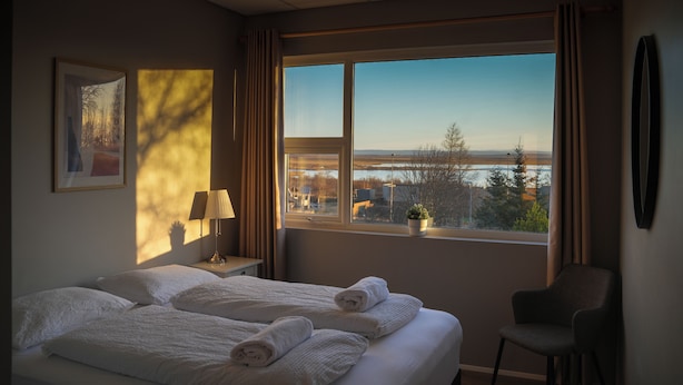 Una habitación de hotel con un ventanal grande, una cama con sábanas blancas, una silla, una mesita de noche con lámpara y un cuadro en la pared.