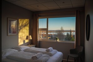 A hotel room with a large window, a bed with white linens, a chair, a nightstand with a lamp, and a framed picture on the wall.
