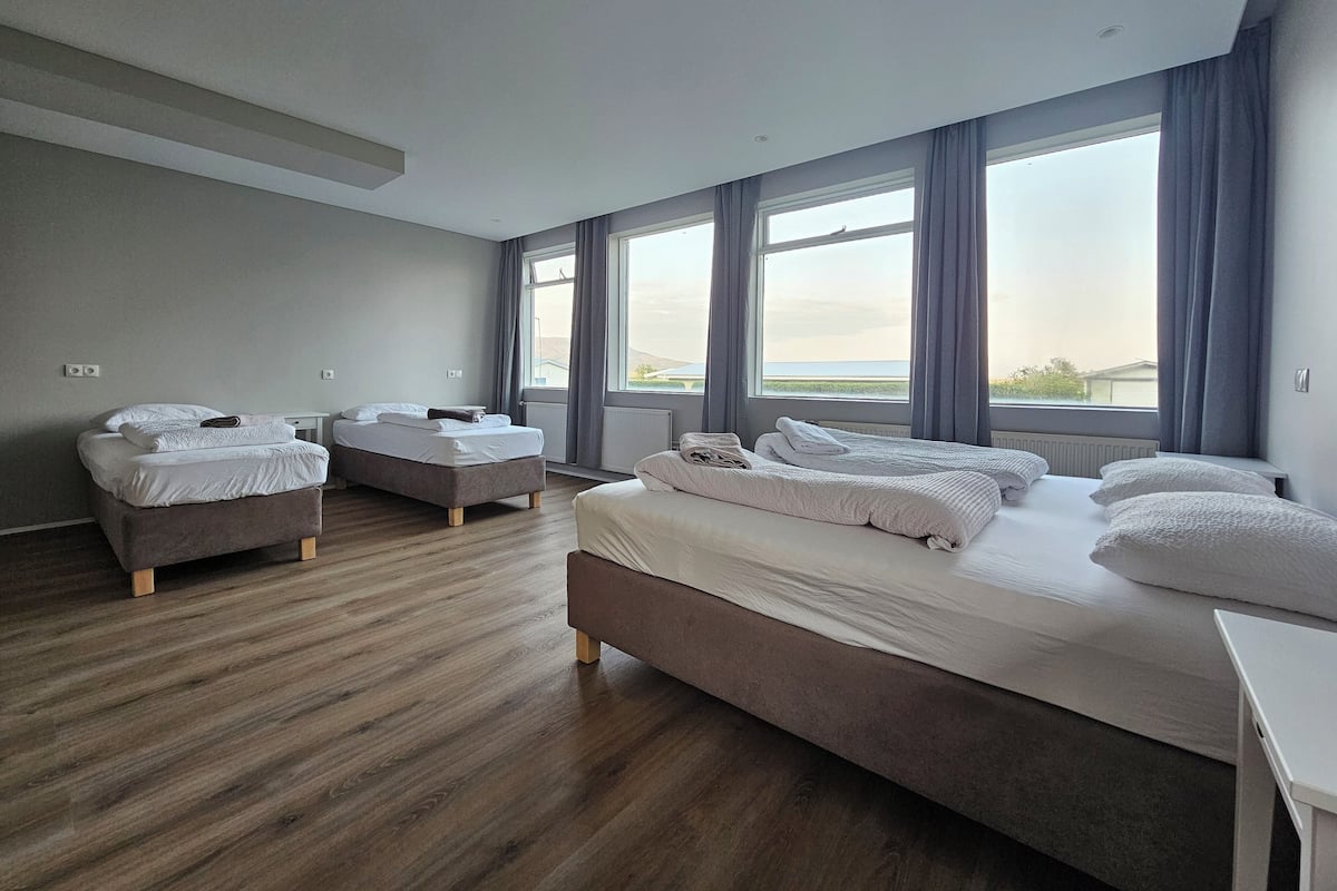 Een moderne hotelkamer met een groot bed, twee kleinere bedden en uitzicht op zee door grote ramen.