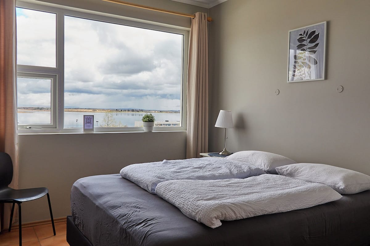 Een slaapkamer met een groot raam, een bed met wit beddengoed, een nachtkastje met een lamp, en een kunstwerk aan de muur.