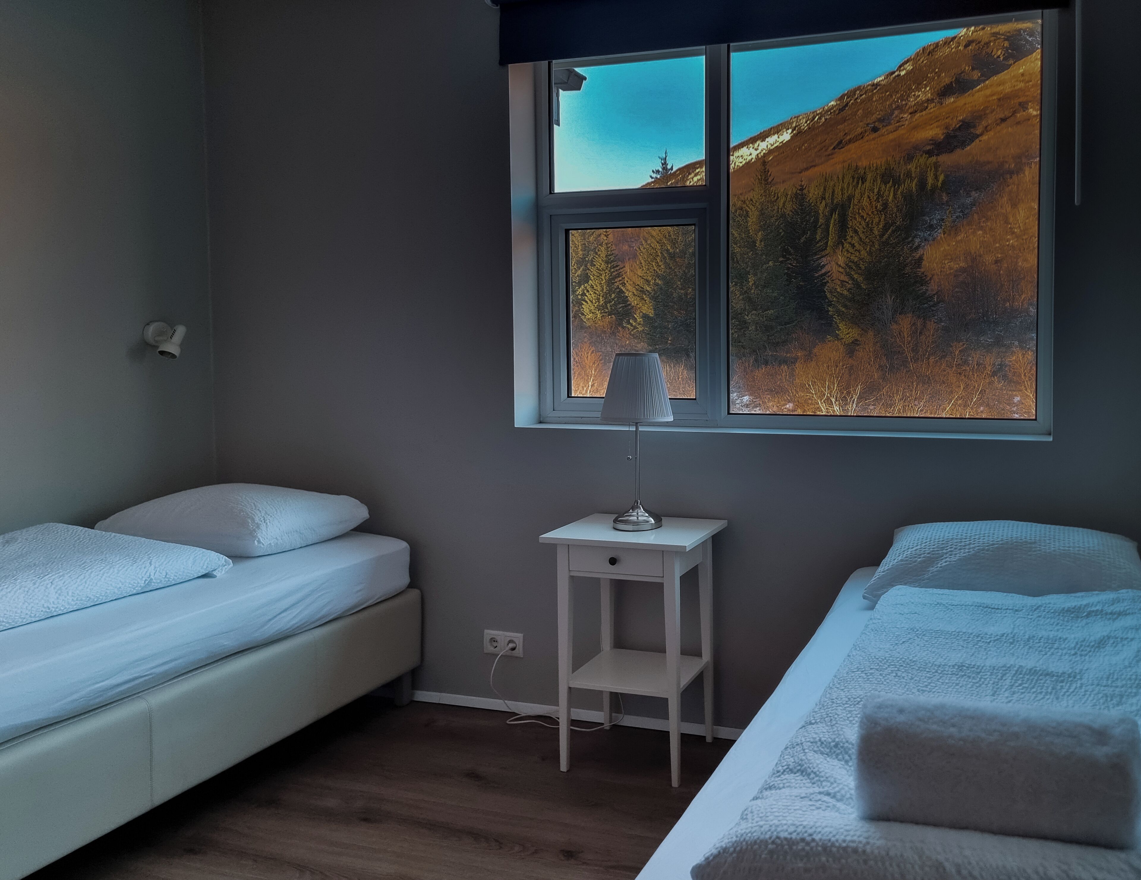 Ein Zimmer mit zwei Einzelbetten, einem kleinen Nachttisch mit Lampe und einem Fenster mit Blick auf eine gebirgige Landschaft.