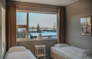 A hotel room with two beds, a window with curtains, a small table with a lamp, and a framed picture on the wall.