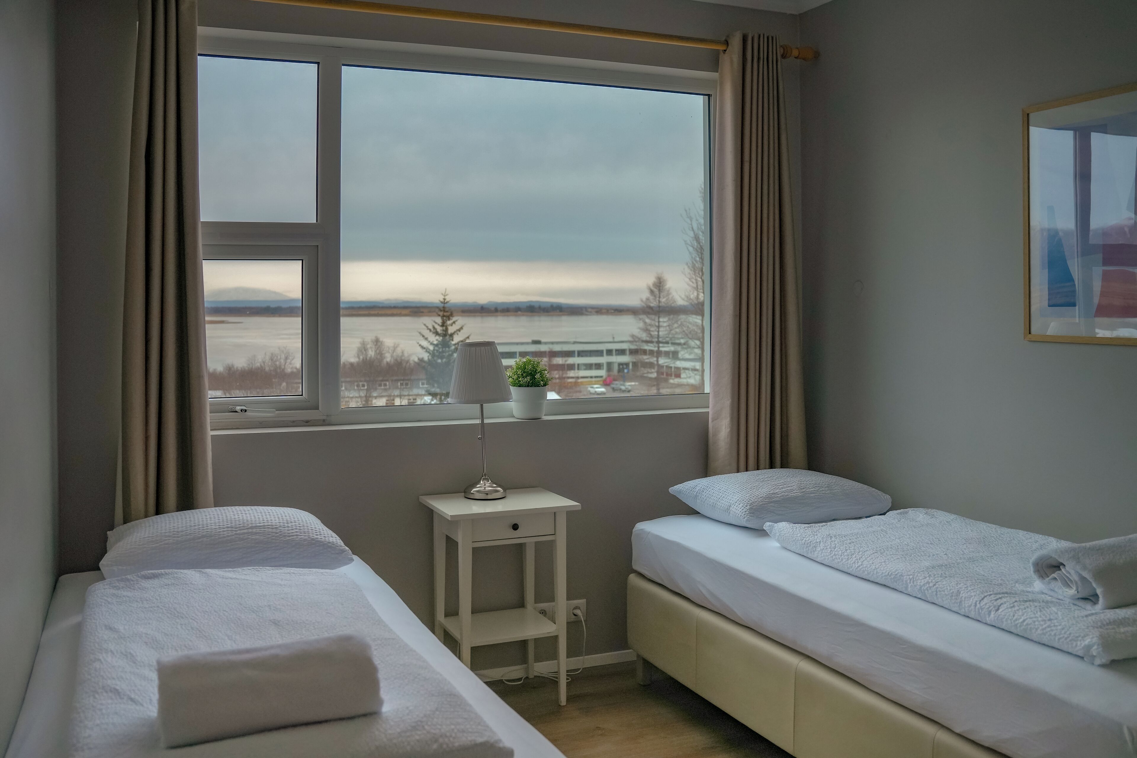 A hotel room with two beds, a window with a view of a body of water and buildings, a small table with a lamp, and a plant.