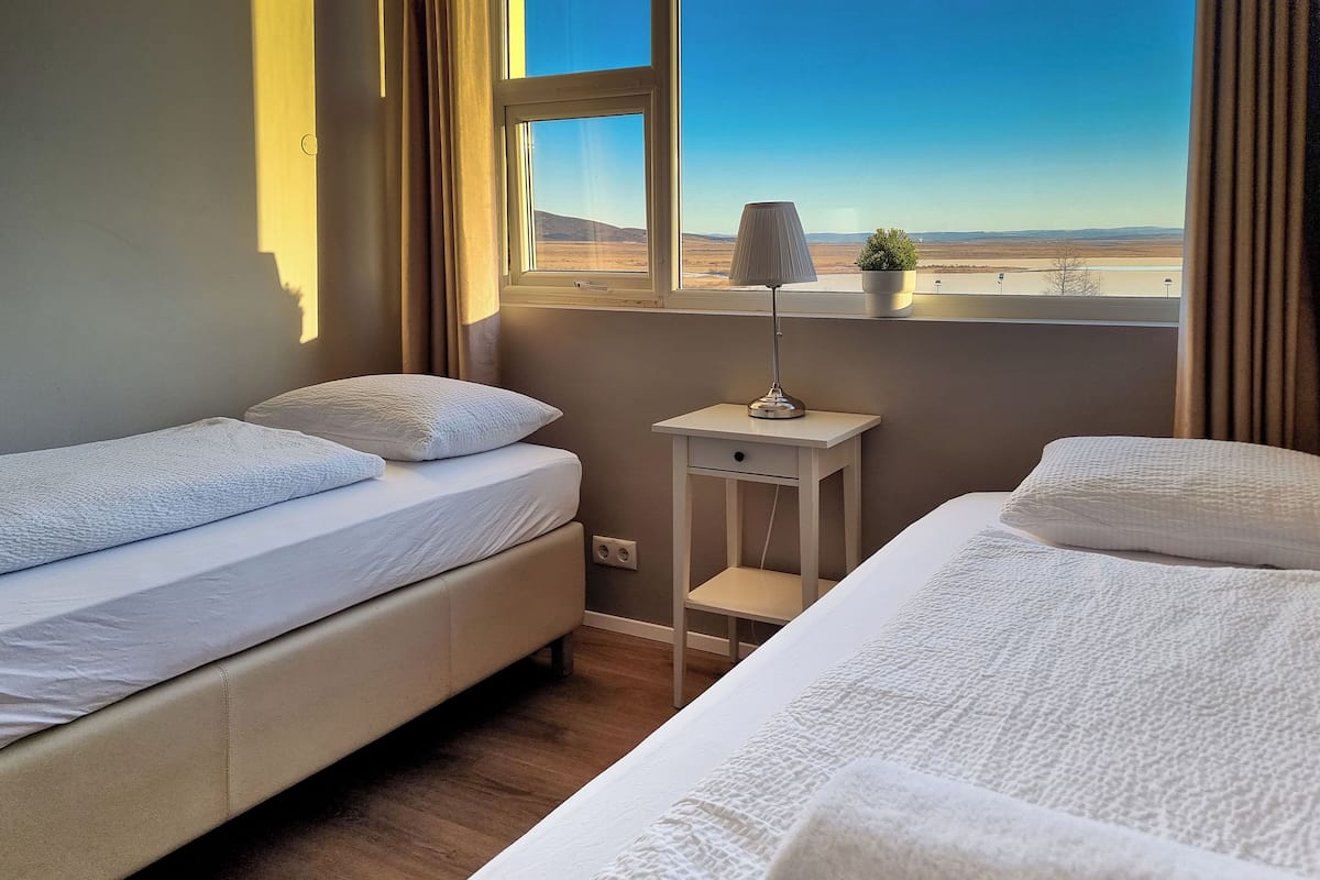 Een slaapkamer met twee bedden, een nachtkastje met een lamp, en uitzicht op een woestijnlandschap door het raam.