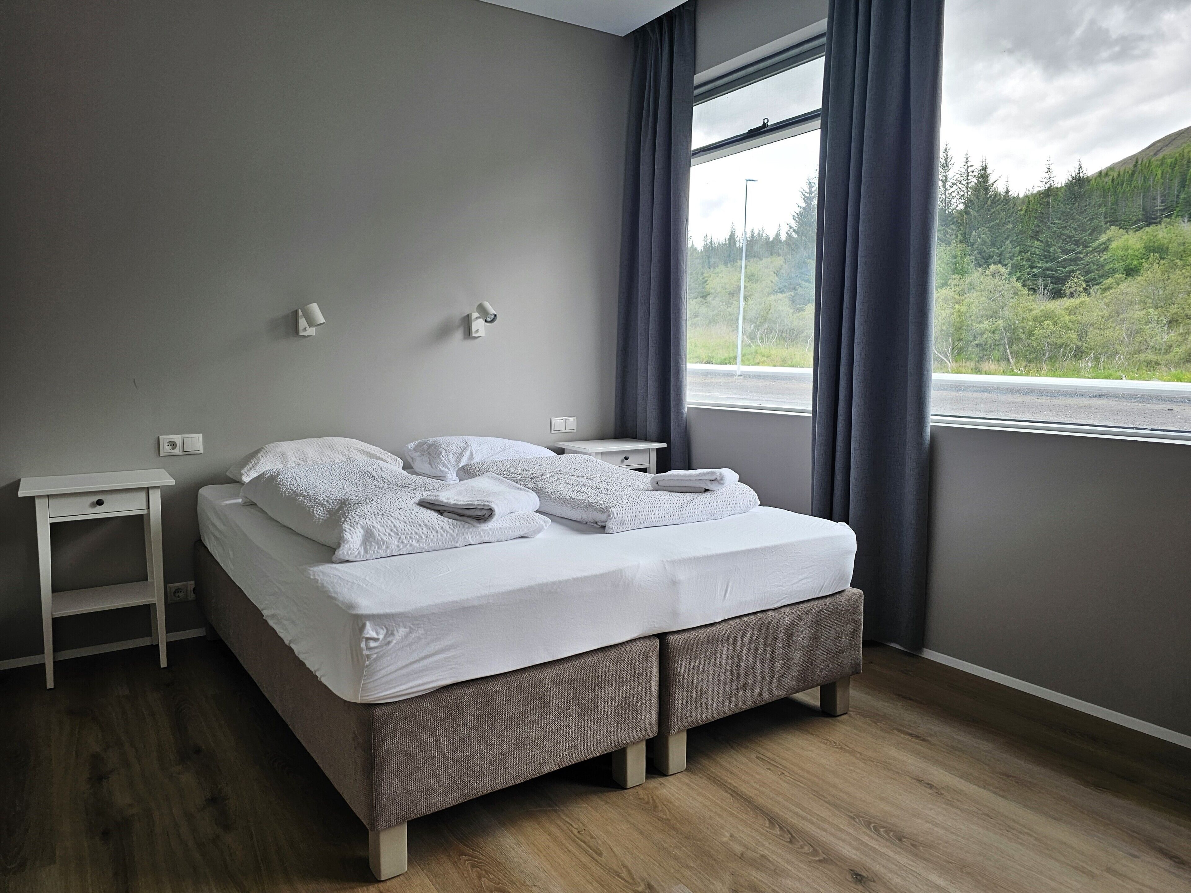 A neatly made bed with white linens, two bedside tables, and a window with a view of a forest and road.