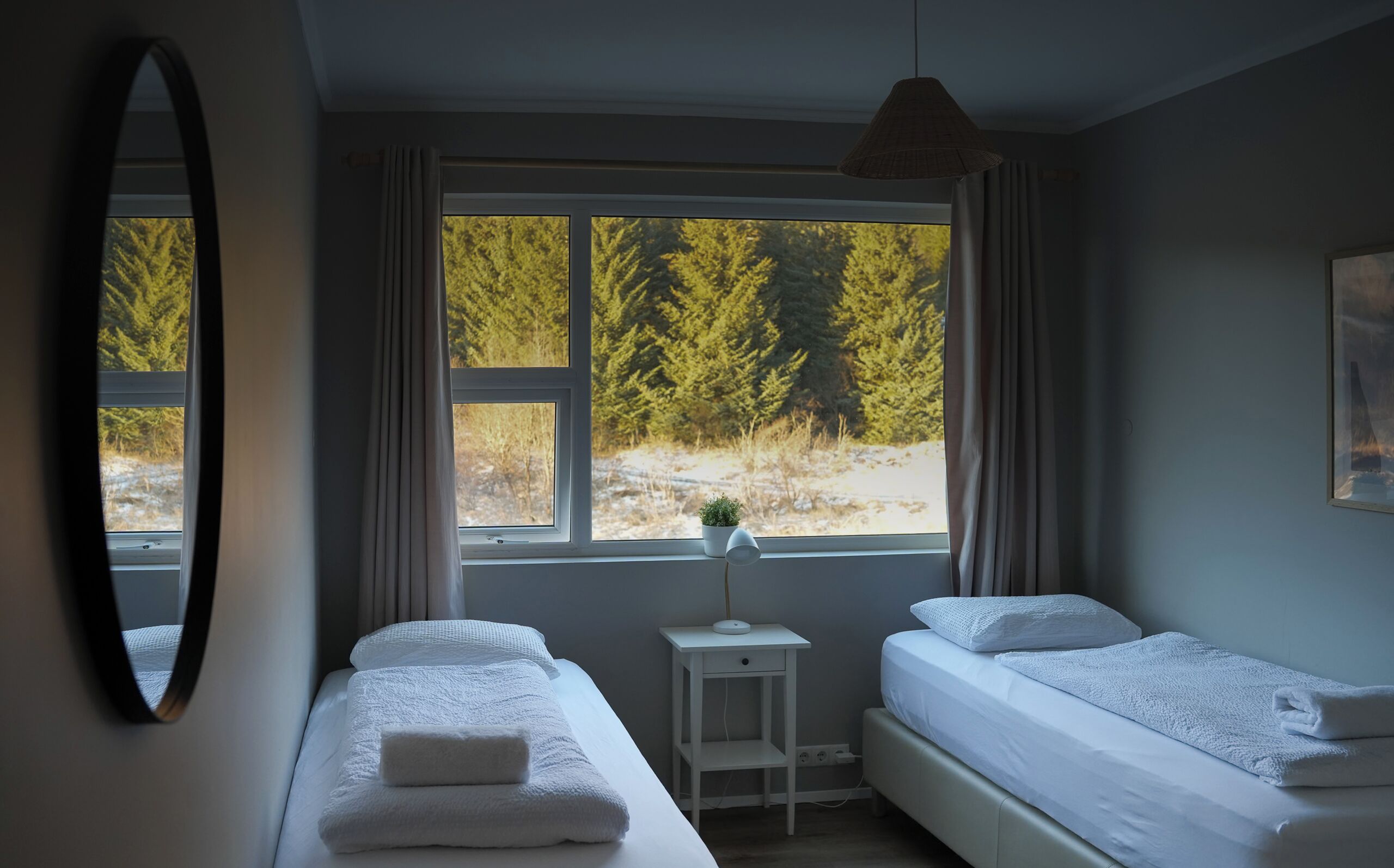 A bedroom with two beds, a small bedside table with a lamp, a window with a view of trees, and a mirror on the wall.