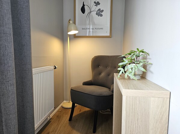 A corner room with a radiator, a grey armchair, a standing floor lamp, and a small table with a potted plant.