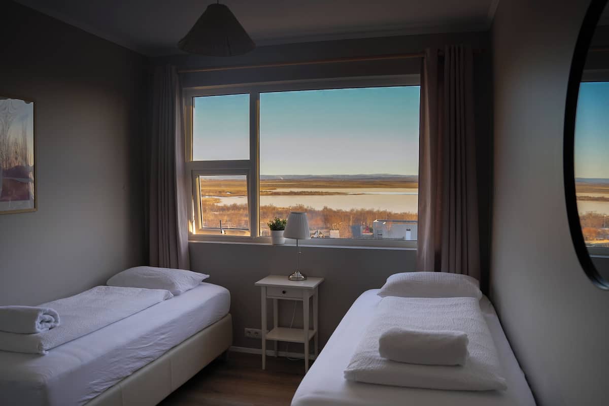 Een kamer met twee bedden, een kleine tafel met een lamp, en een groot raam met uitzicht op een vlak landschap.