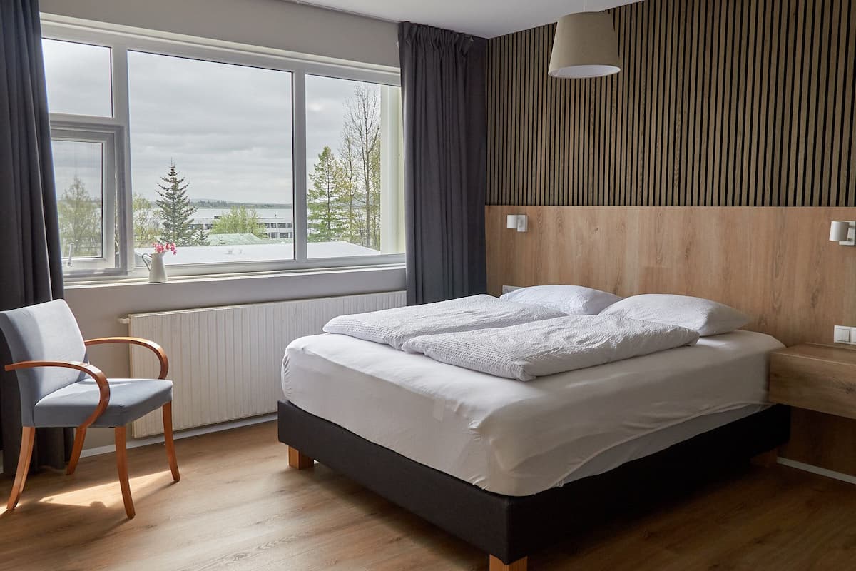 Een slaapkamer met een groot raam, een bed met witte lakens, een houten hoofdeinde en een stoel.