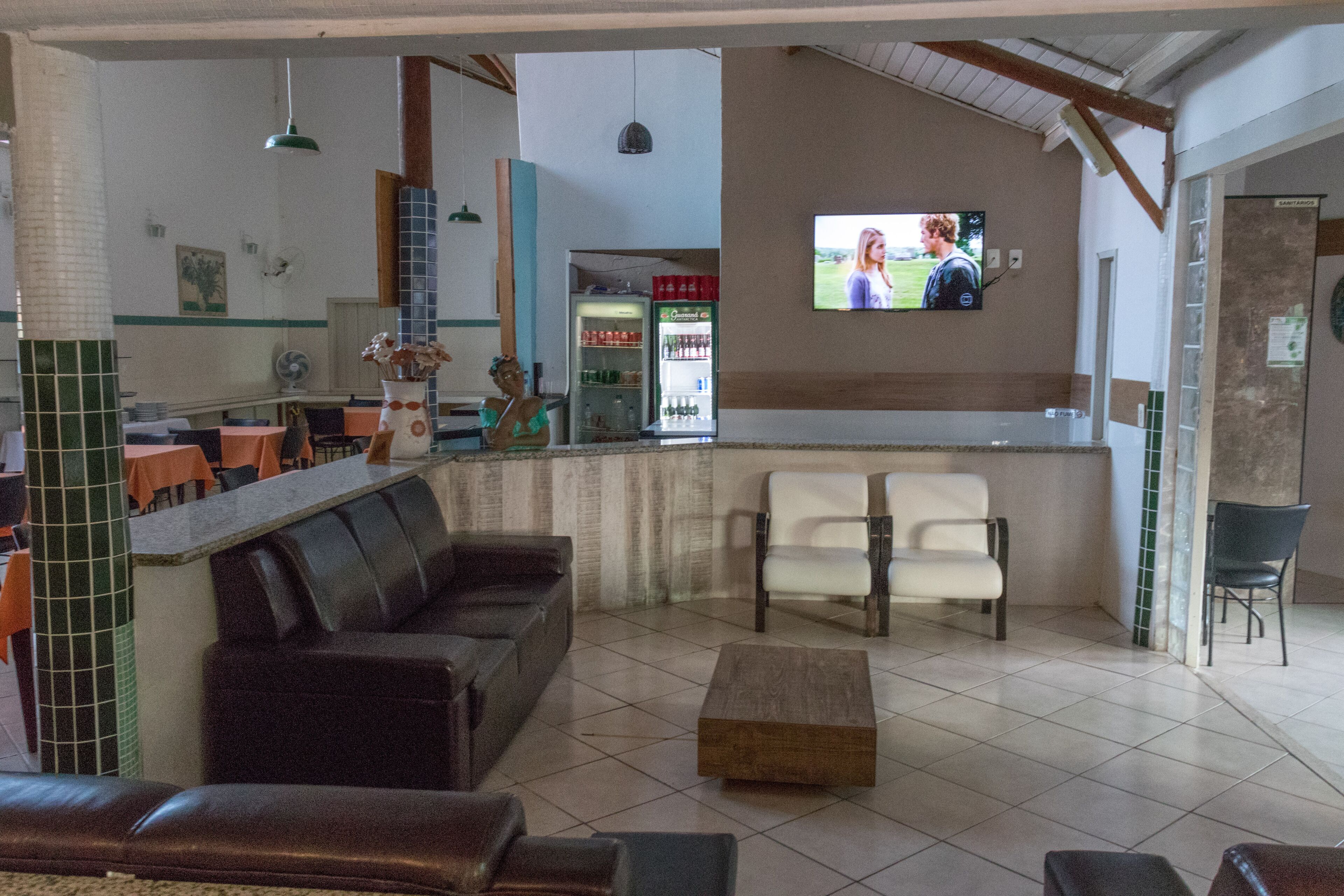 Restaurante com área de bar, mesas e cadeiras, e uma tela de televisão.