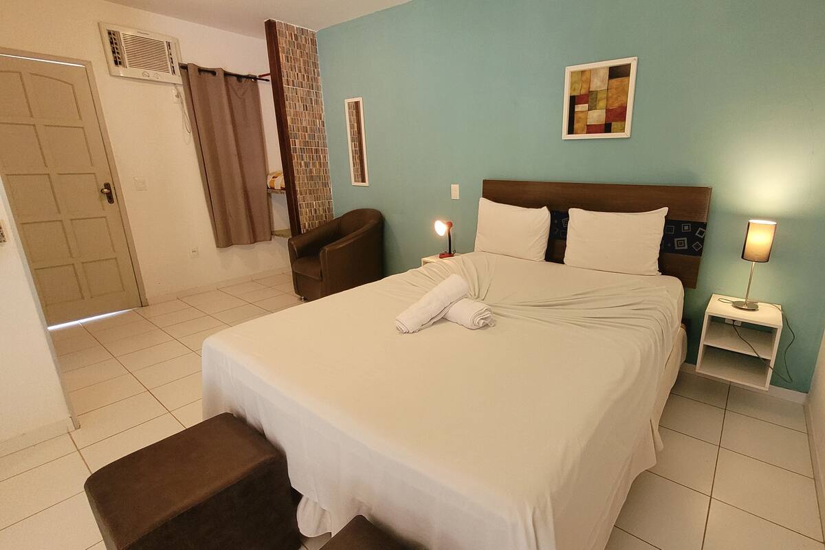 A hotel room with a bed, a chair, a nightstand, a lamp, a window, and a door.