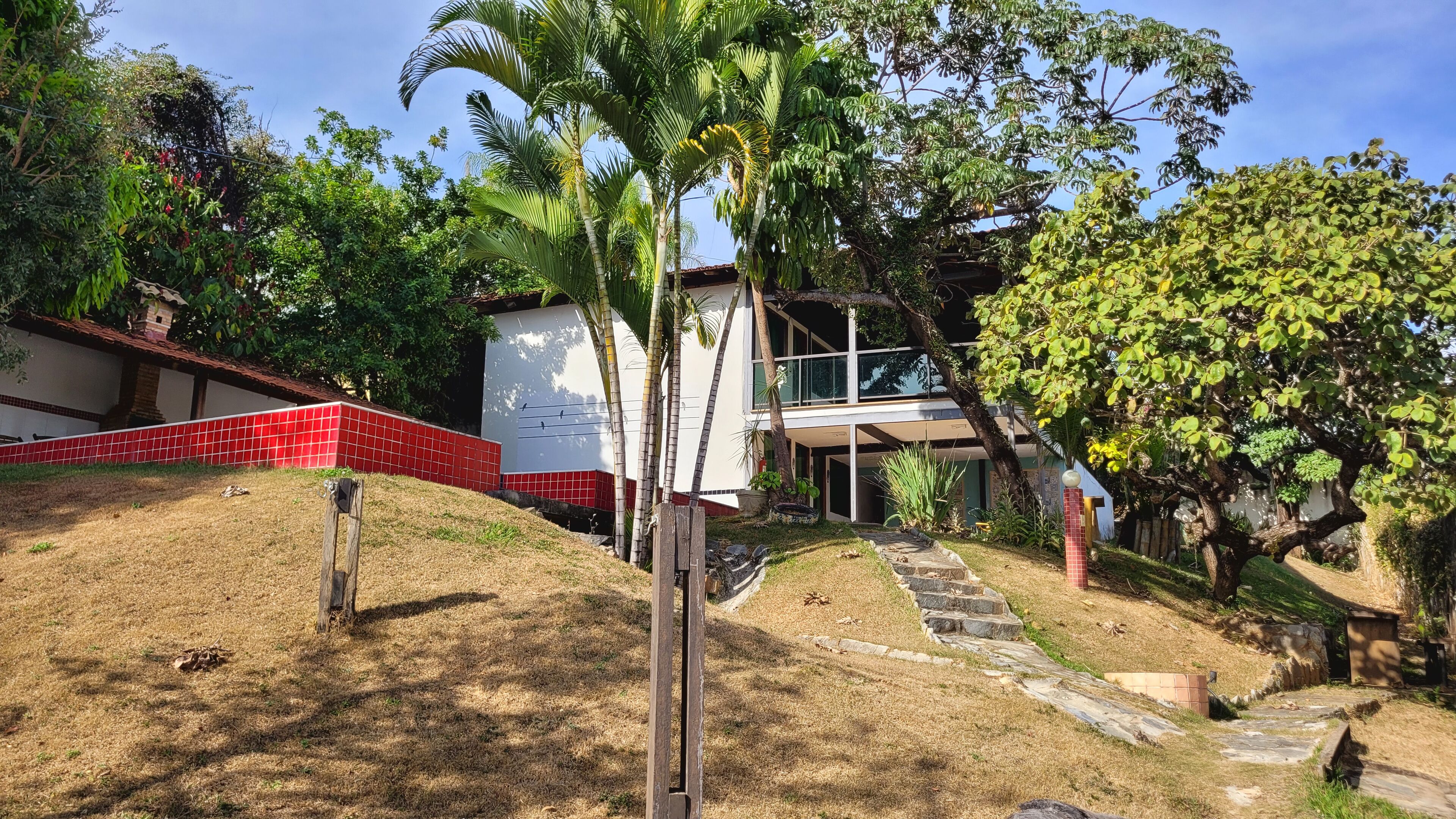 Un bâtiment à l’extérieur recouvert de tuiles rouges, un chemin de pierre menant à l’entrée, et une variété d’arbres et d’arbustes dans les environs.
