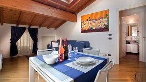 A dining area with a table set for two, a painting on the wall, and a view of the kitchen through an open door. - Verona For Rent La Colombina (Verona)