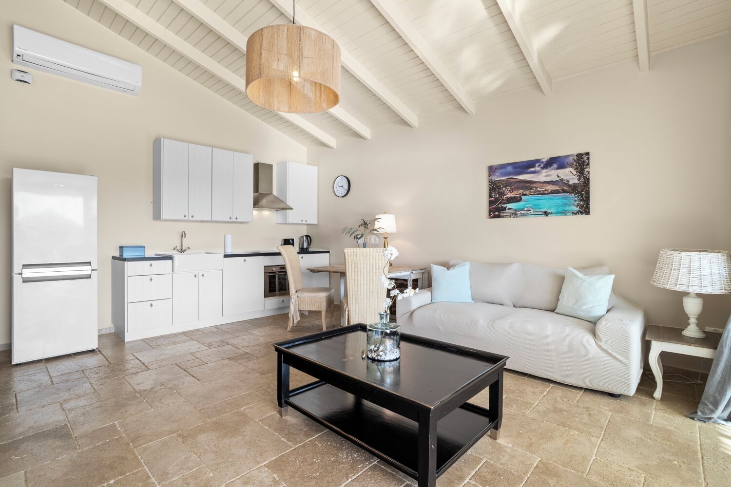 Una cocina moderna con armarios blancos, un comedor con mesa y sillas, una sala de estar con sofá, y un cuadro en la pared.