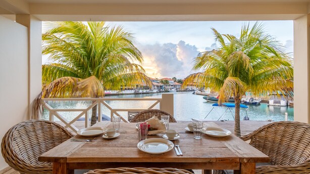 A wooden table set for dining with wicker chairs, overlooking a marina with boats and palm trees. - Caribbean Lofts Bonaire (Kralendijk)
