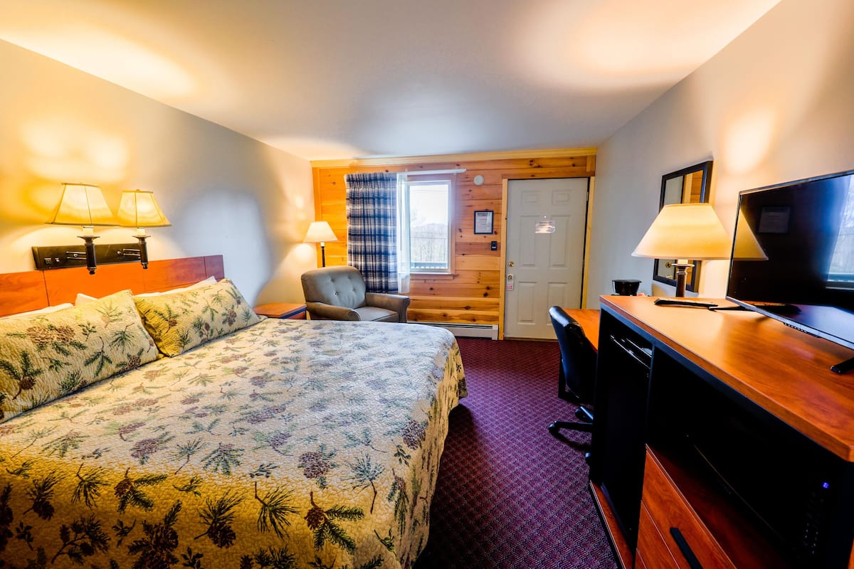 Quarto de hotel com cama, escrivaninha, cadeira, televisão e janela com cortinas.