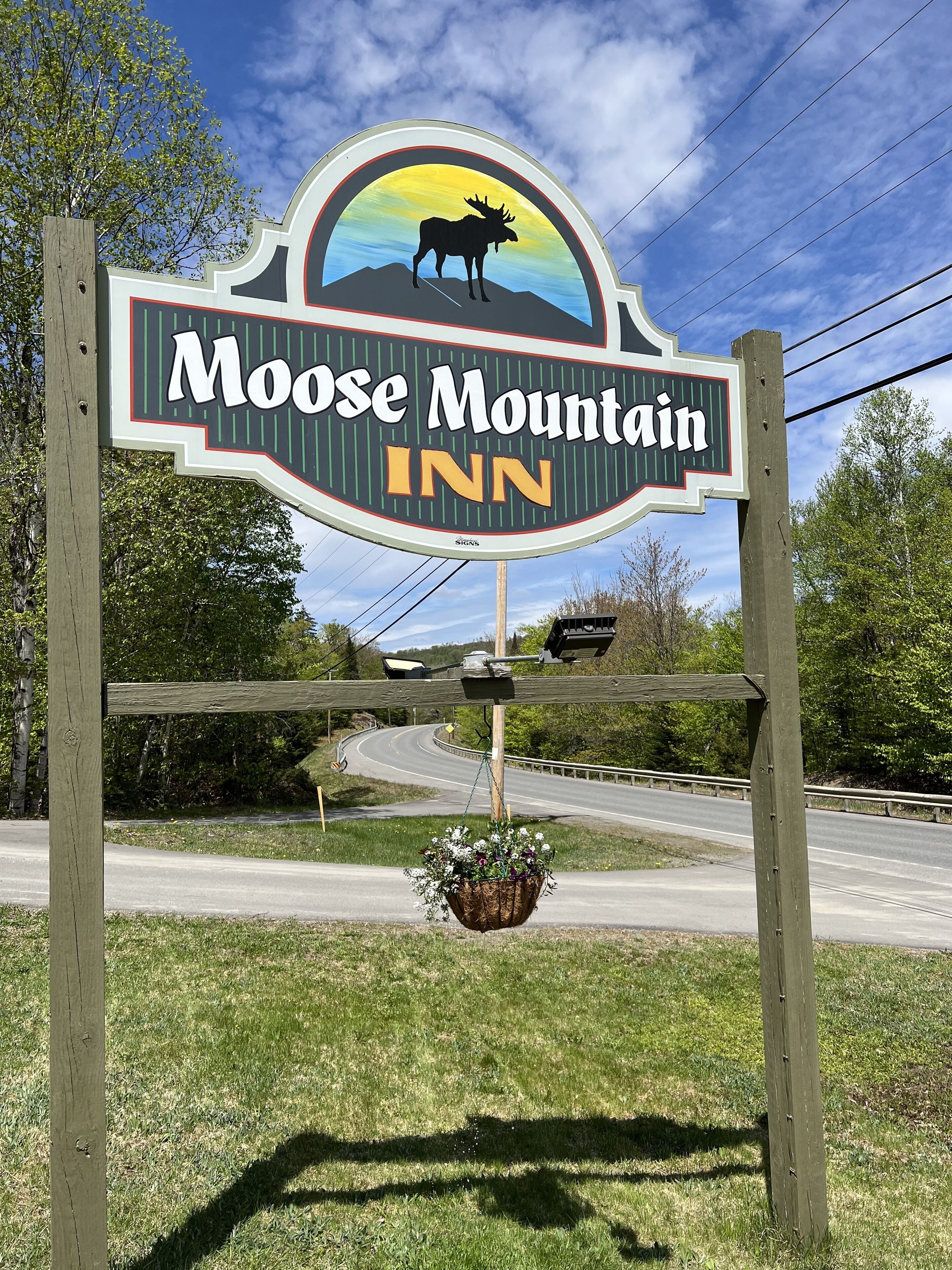 A sign for Moose Mountain Inn with a moose silhouette, a sunset background, and a hanging flower basket.