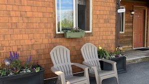 A brick building with a window, two Adirondack chairs, and a planter with flowers.