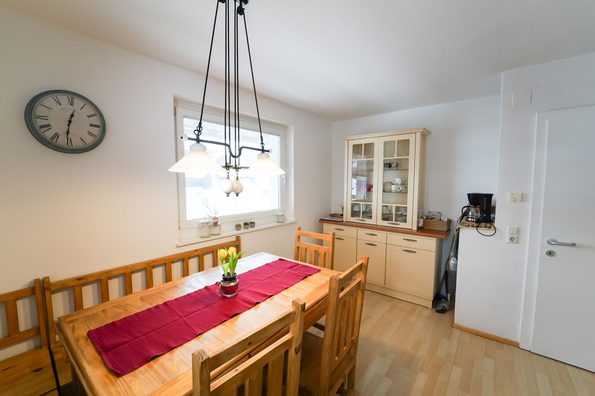 A dining area with a wooden table, chairs, a red table runner, and a clock on the wall.
