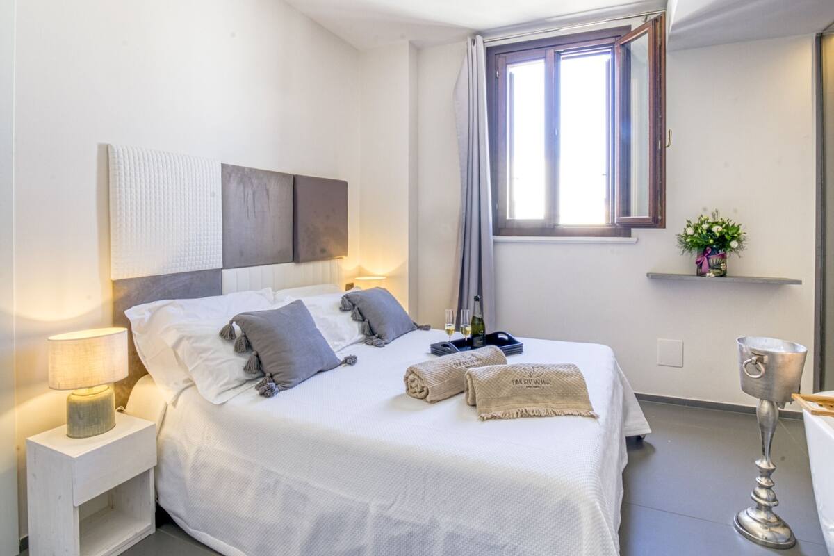 A modern bedroom with a large bed, bedside tables, a window with curtains, and a small shelf with a plant.