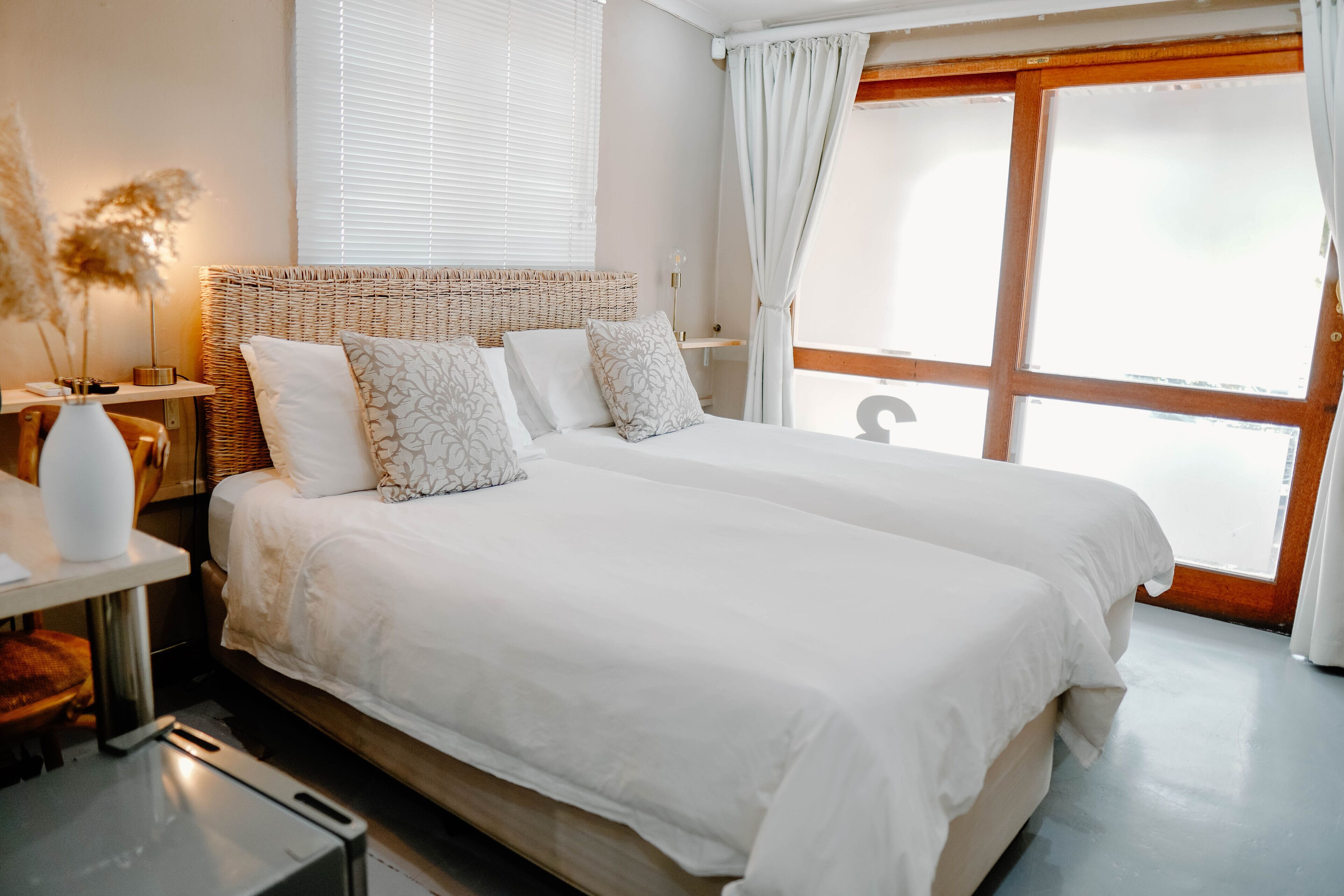 A double bed with white linens and patterned pillows, a bedside table with a lamp, and a window with curtains.