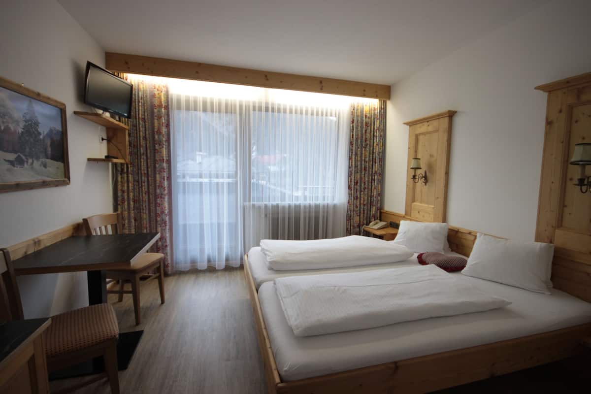 A hotel room with a wooden bed, a desk with a chair, a television on a shelf, and a window with sheer curtains.