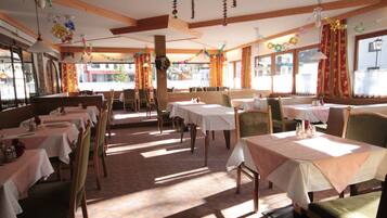 Ein Restaurant mit mehreren Tischen und Stühlen, Holzbalken an der Decke und großen Fenstern mit Vorhängen.