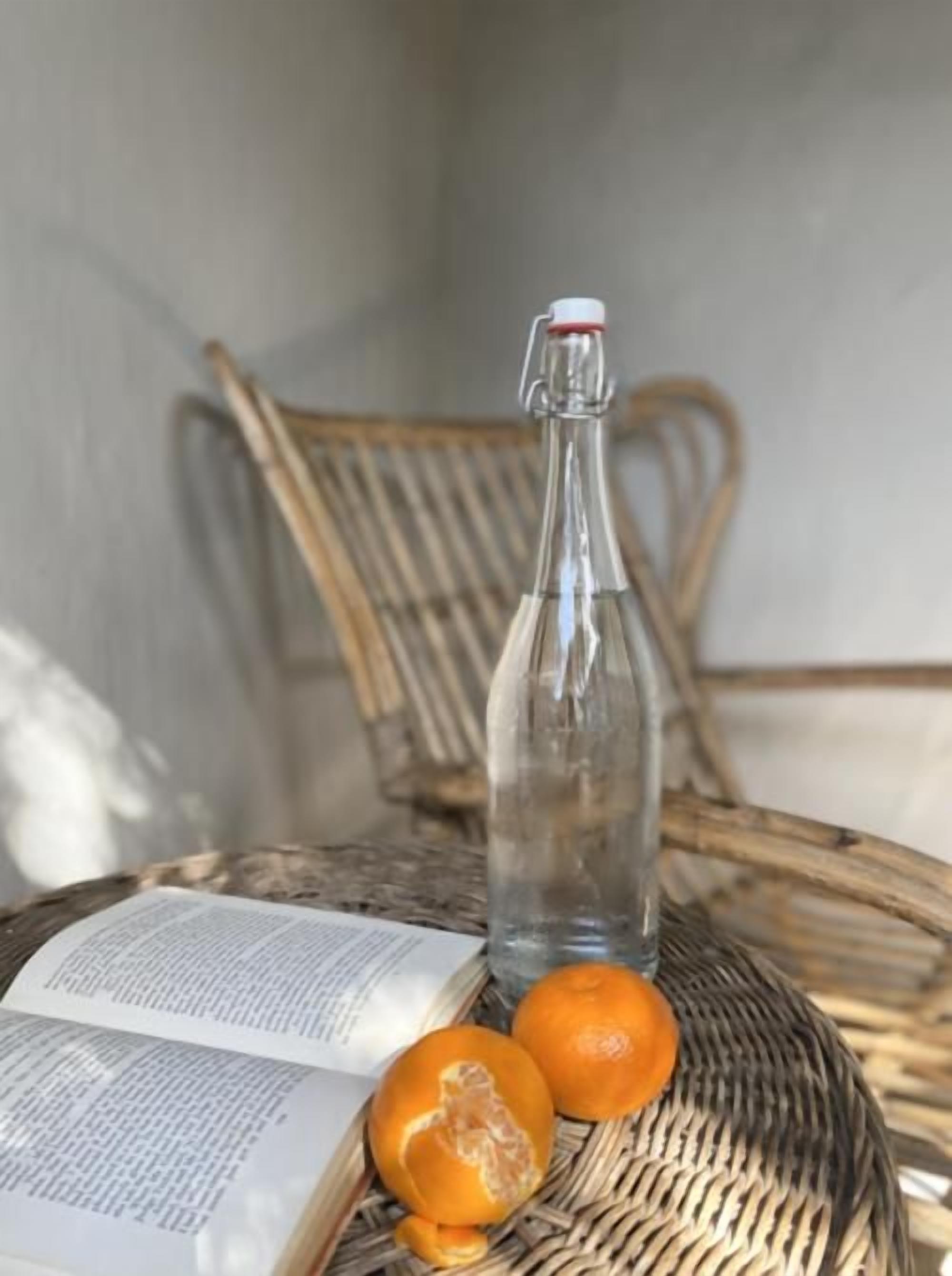 Una sedia in vimini, una bottiglia di vetro trasparente e un libro aperto con un'arancia accanto.