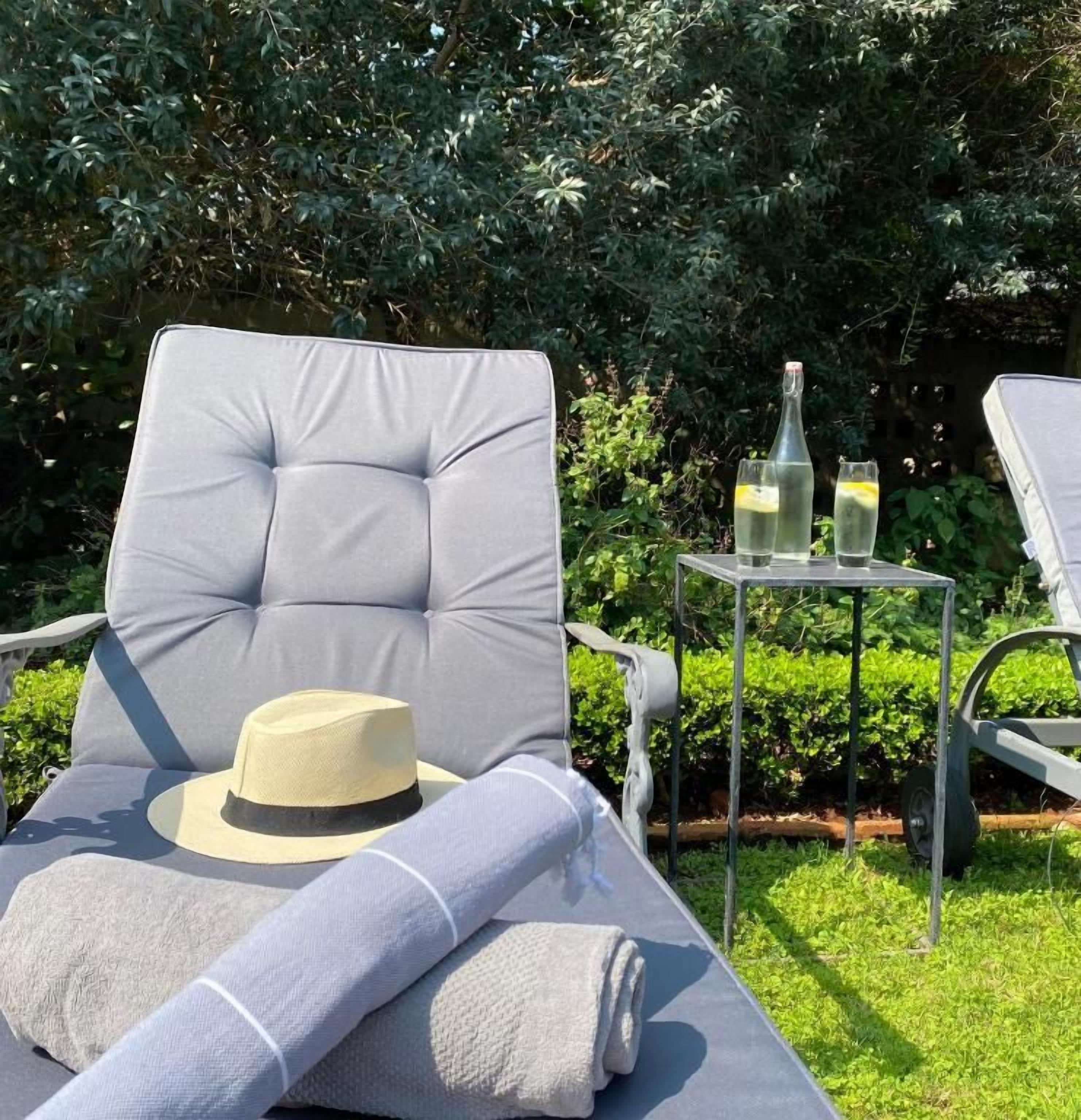 A lounge chair with a cushion, a rolled towel, and a hat on the seat. There is a small table with two glasses and a bottle.