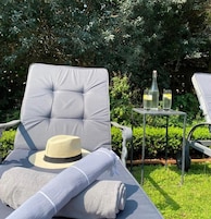A lounge chair with a cushion, a rolled towel, and a hat on the seat. There is a small table with two glasses and a bottle.