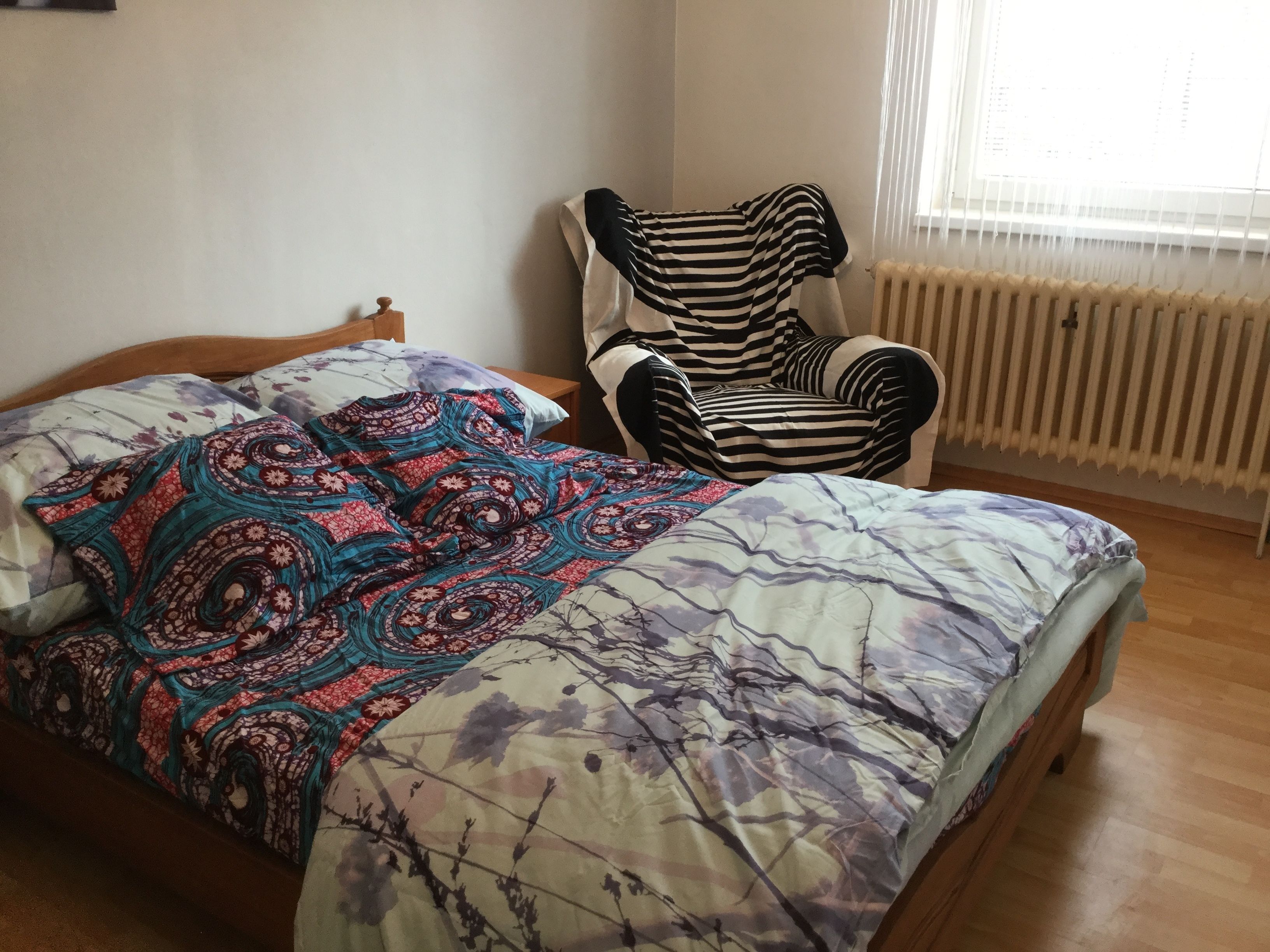 A bed with a floral-patterned bedspread, a striped armchair, and a radiator in a room with wooden flooring.