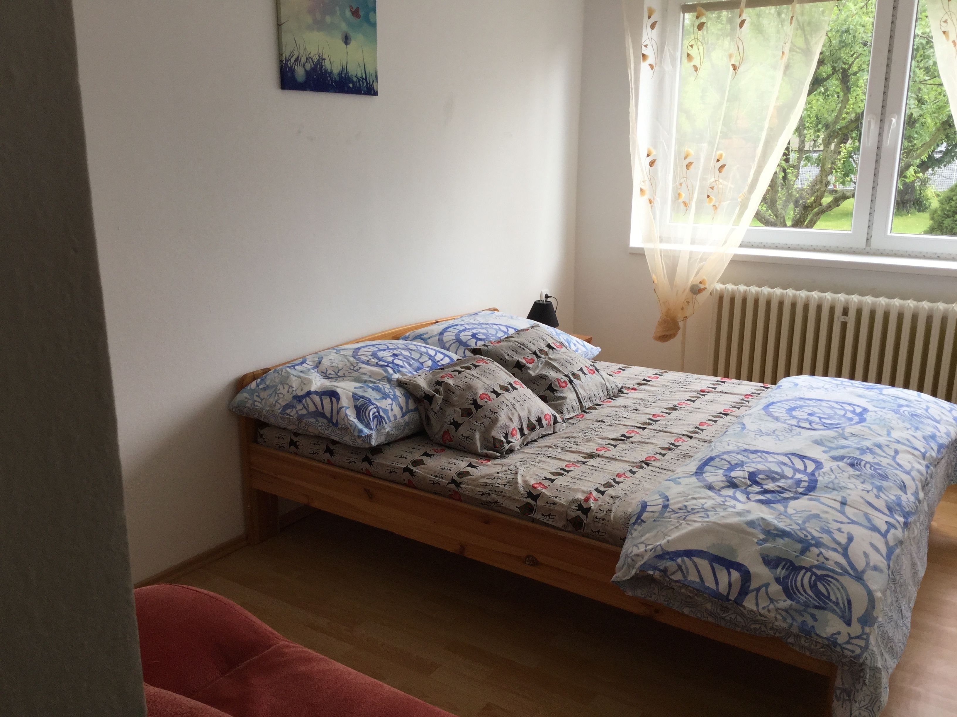 A bedroom with a wooden bed, patterned bedding, a small bedside table, and a window with sheer curtains.