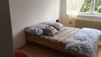 A bedroom with a wooden bed, patterned bedding, a small bedside table, and a window with sheer curtains.