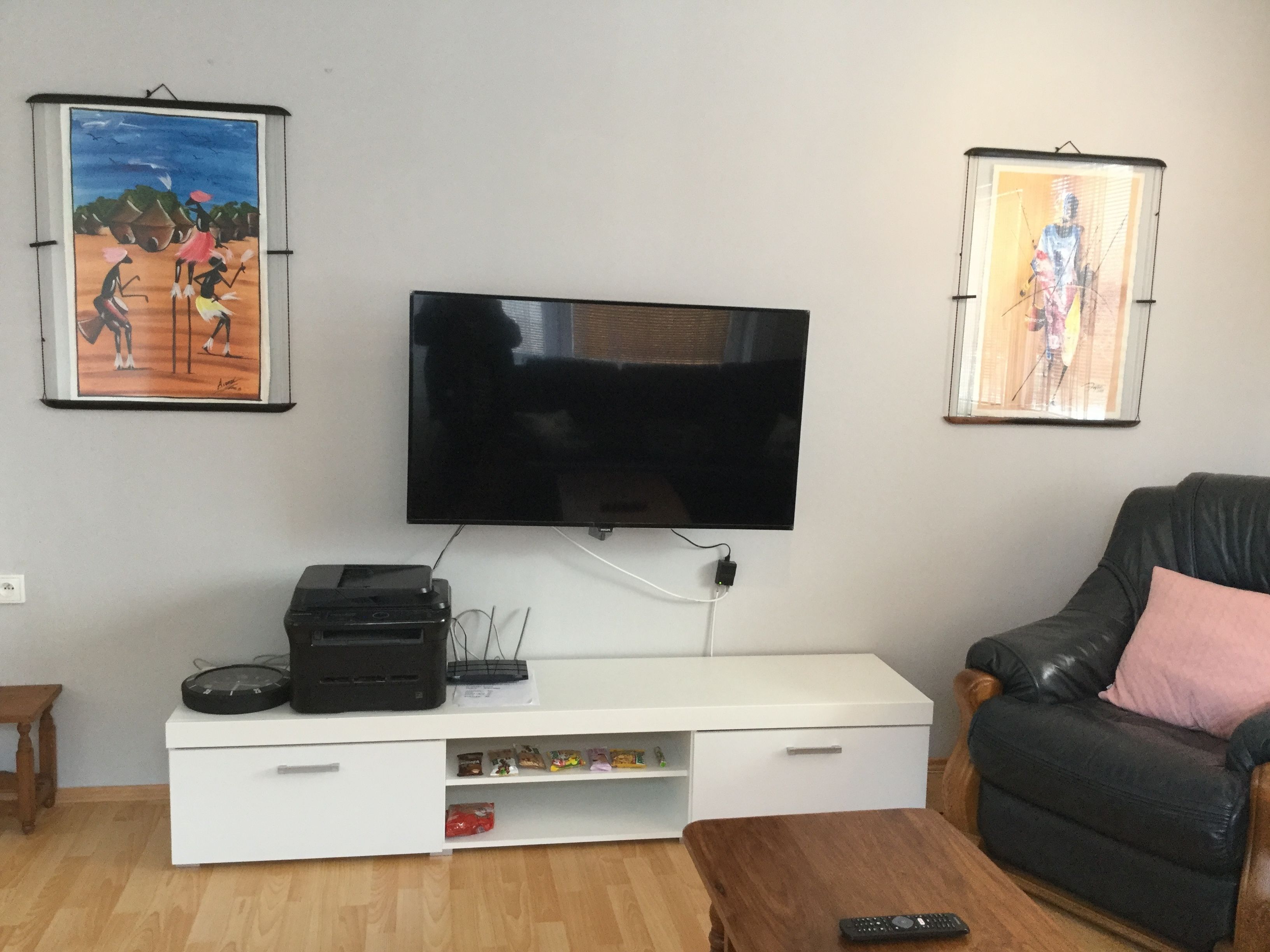 A living room with a flat-screen TV mounted on the wall, a white entertainment unit with a printer, a leather sofa with a pink pillow, and two framed artworks on the walls.