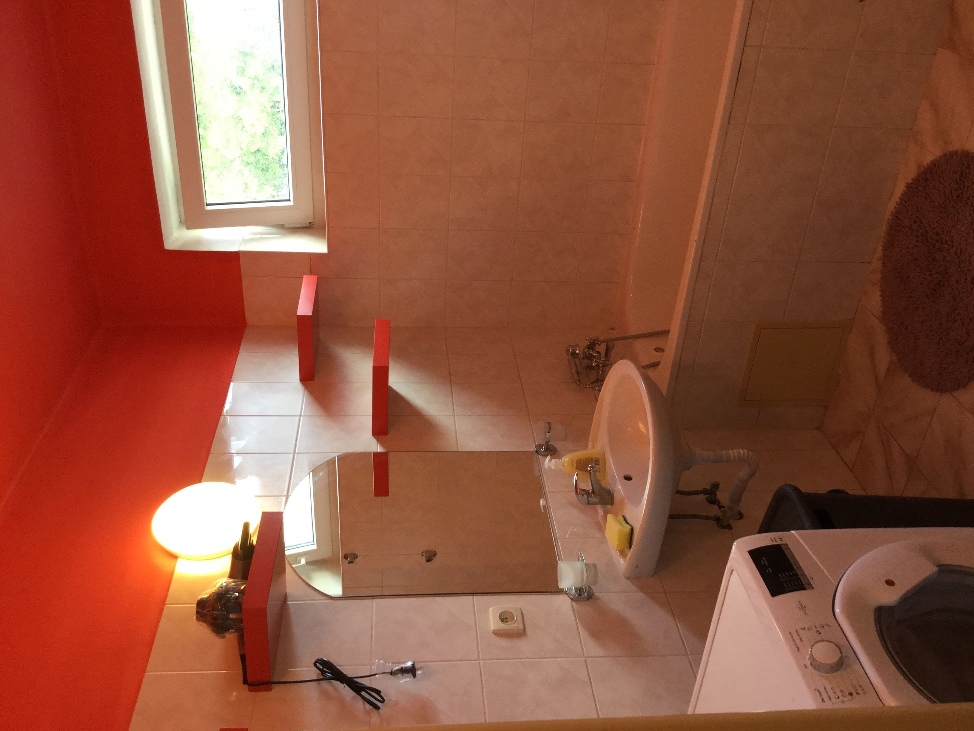 A bathroom with a white sink, toilet, and a washing machine. There is a window providing natural light and a red wall.