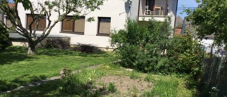 A two-story house with a balcony, surrounded by a garden with various plants and a small tree.