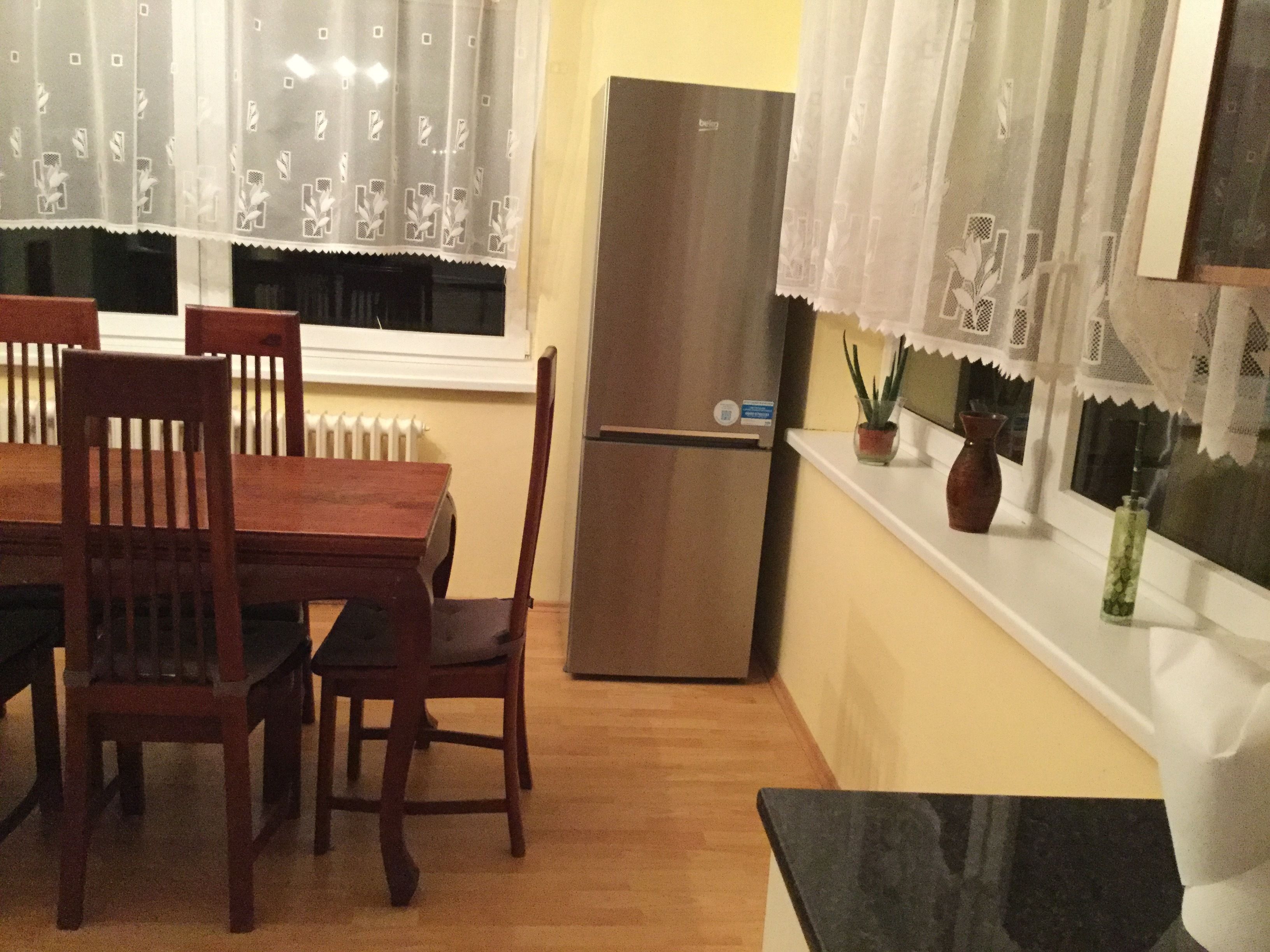 A dining area with a wooden table and chairs, a refrigerator, and a window with curtains.