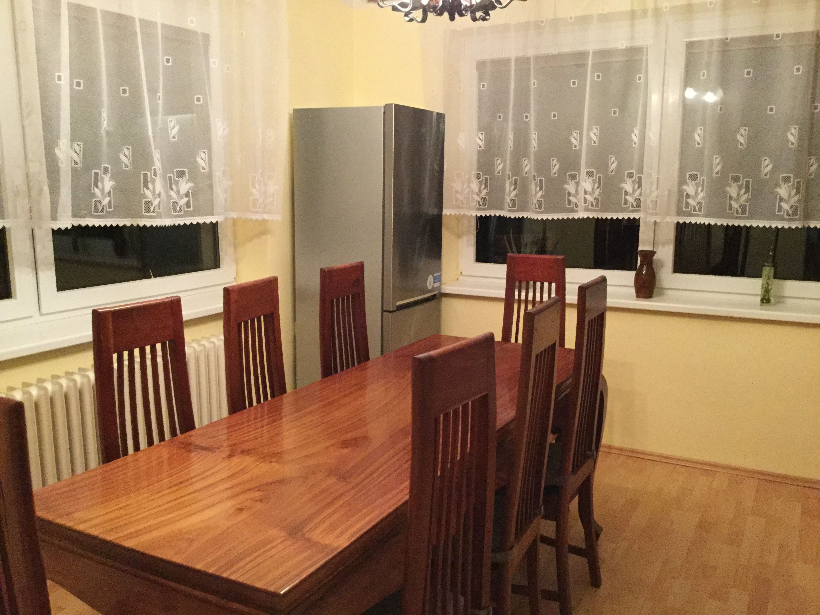 A dining area with a wooden table and six chairs, a refrigerator, and two windows with patterned curtains.