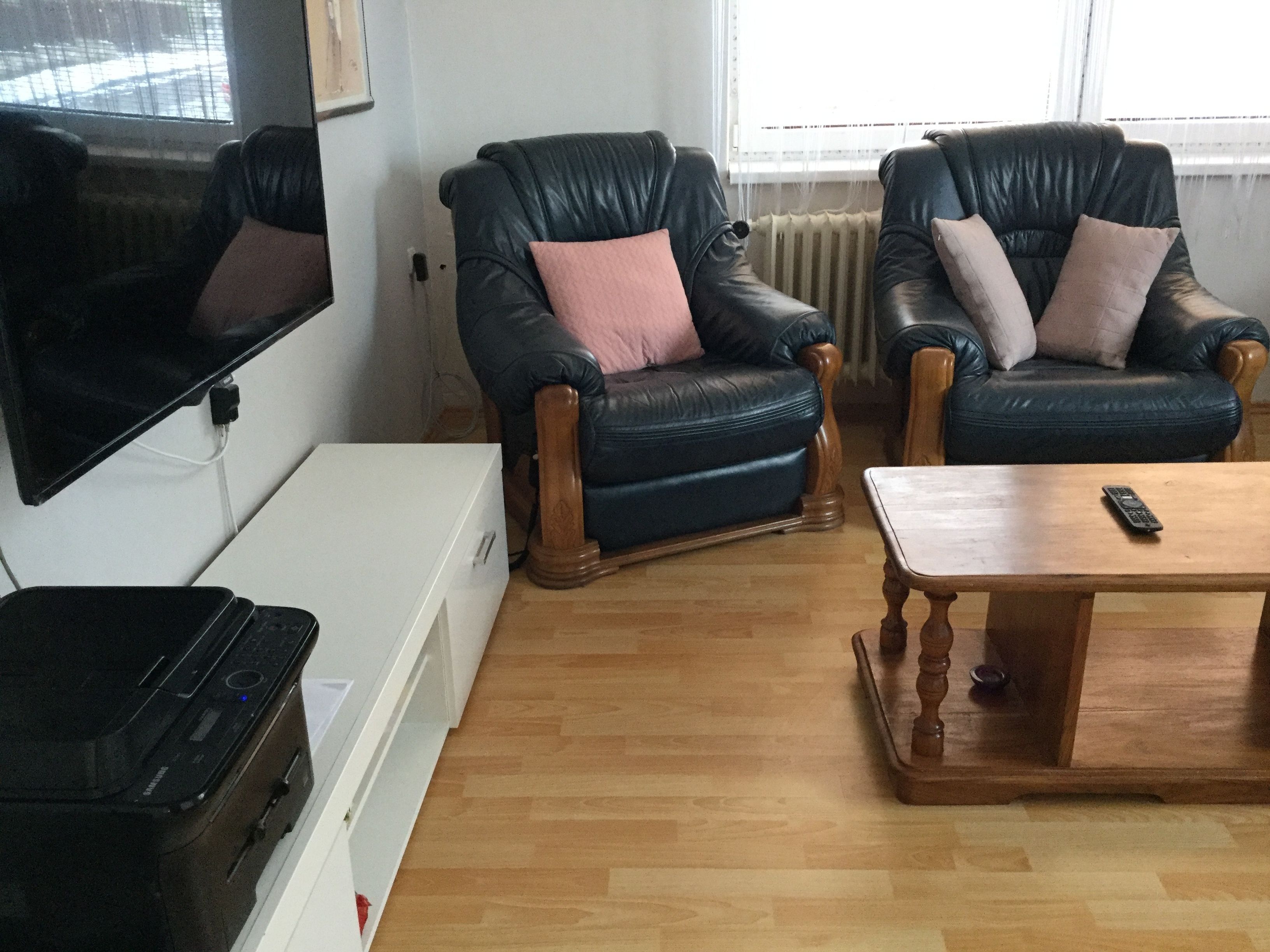 A living room with two leather armchairs, a wooden coffee table, a TV mounted on the wall, and a white cabinet.