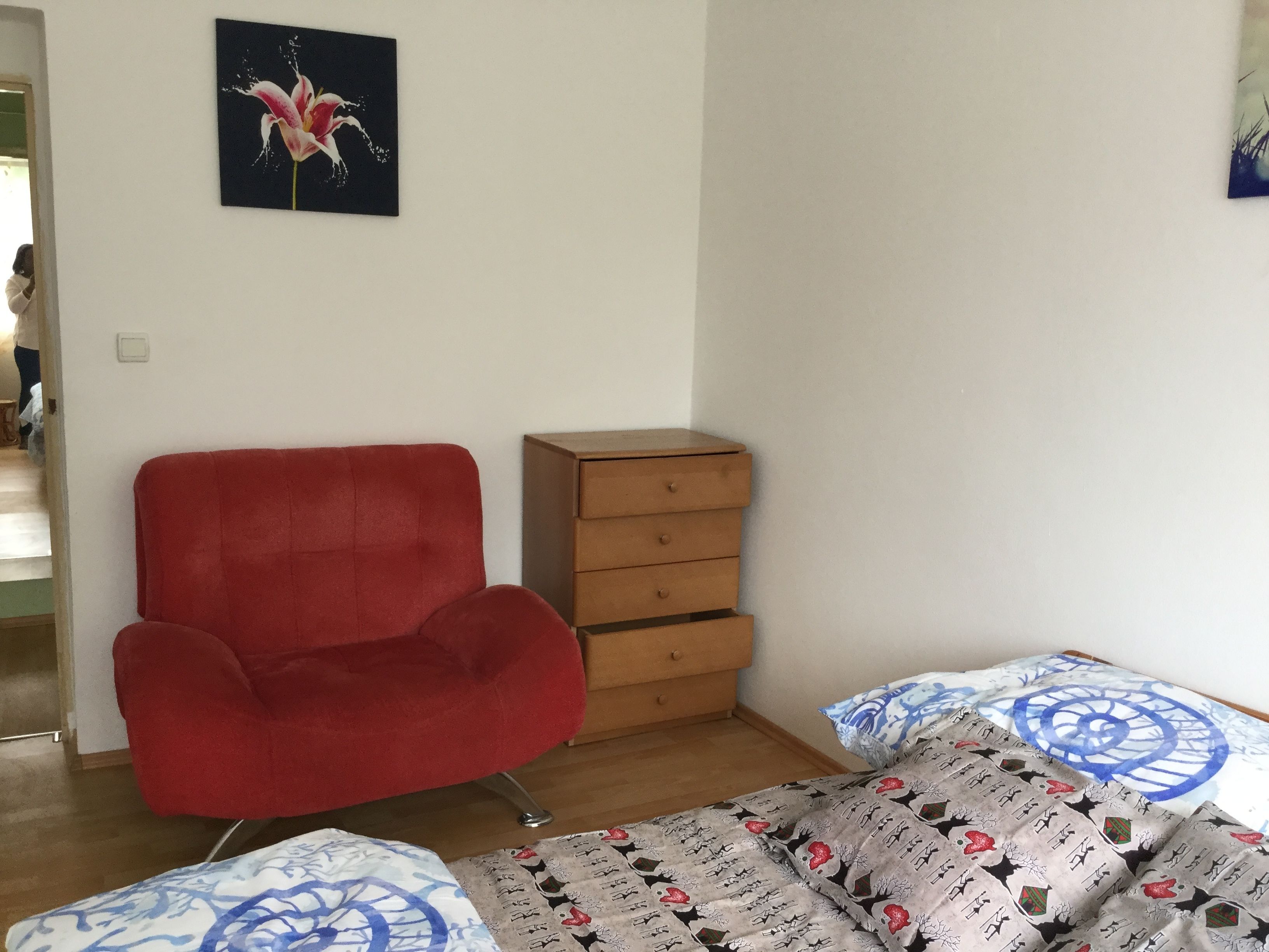 A room with a red armchair, a wooden chest of drawers, and a bed with patterned bedding.