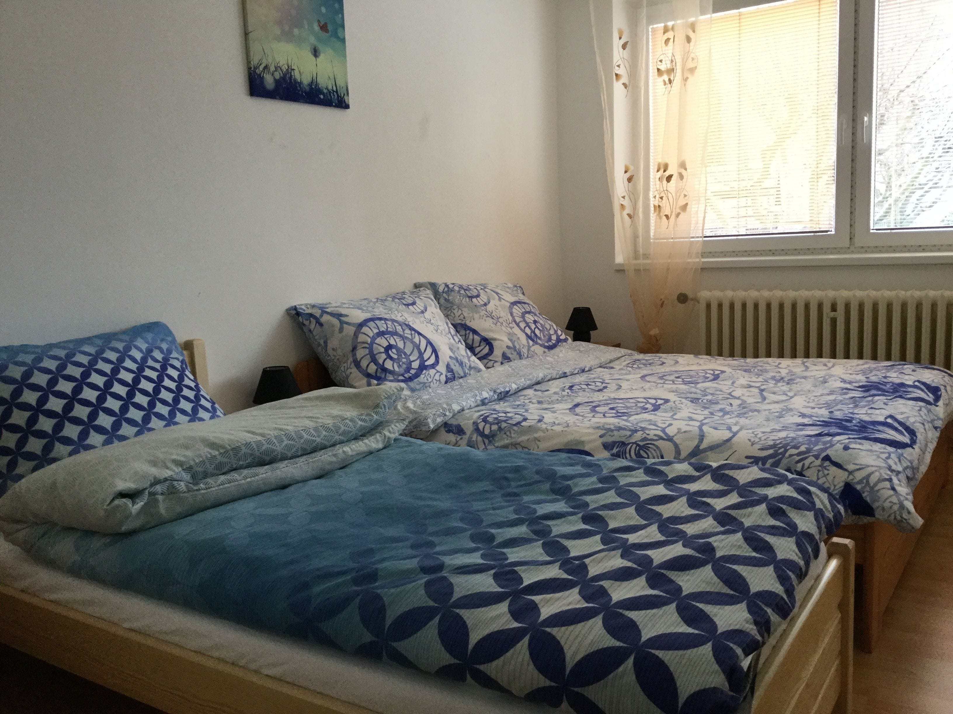 A bedroom with two beds, each with patterned bedding. There is a window with sheer curtains and a radiator below it.
