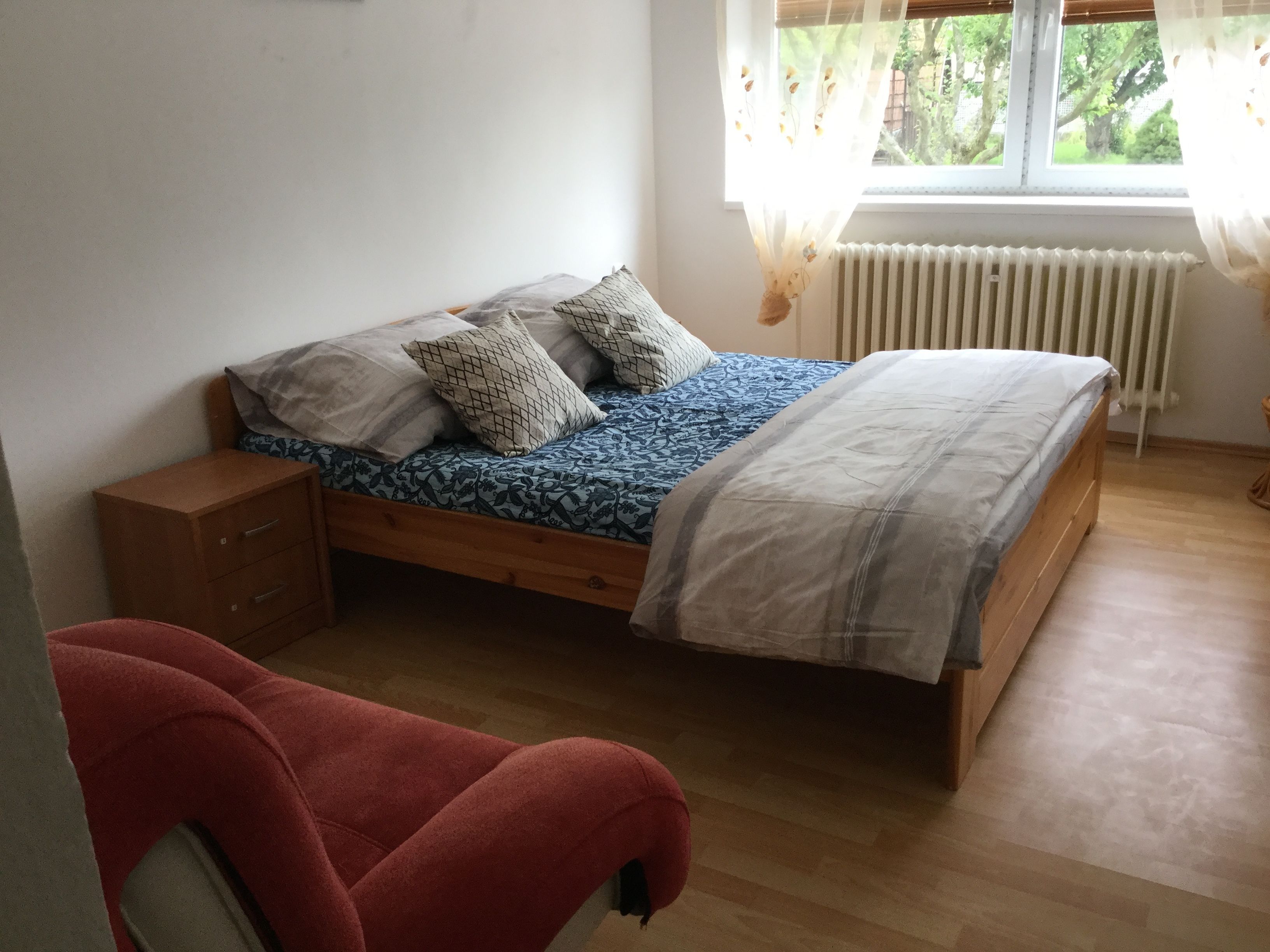 A bedroom with a wooden bed, a nightstand, a red chair, and a window with a view of trees.
