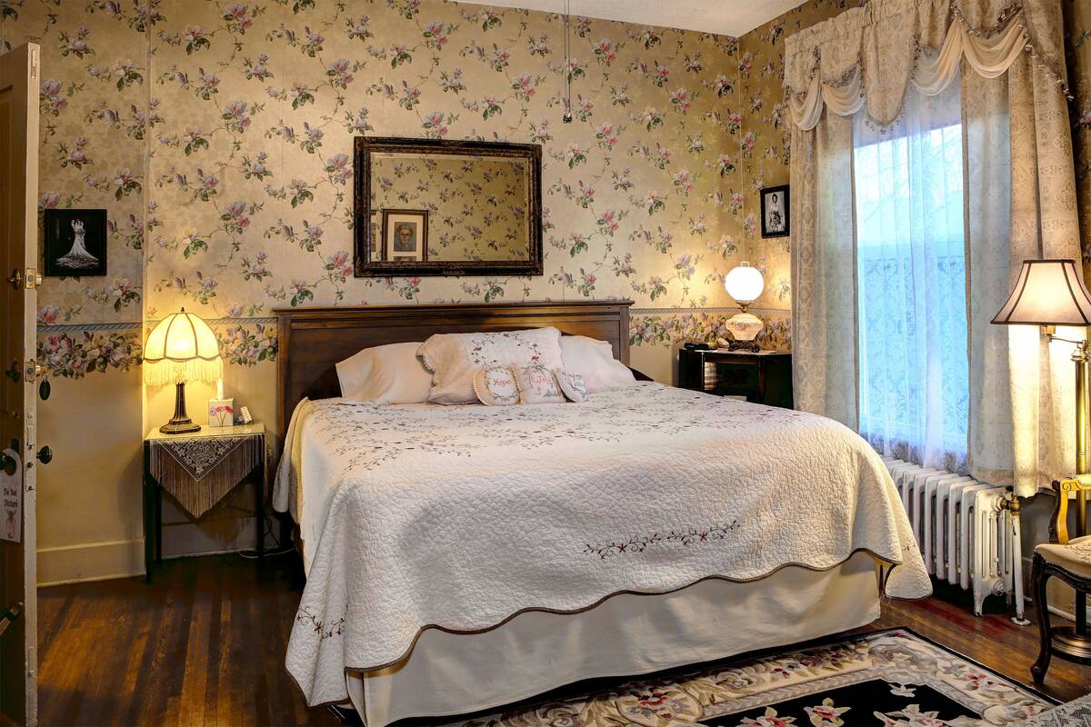 A bedroom with floral wallpaper, a large bed with a white quilt, two bedside tables with lamps, and a radiator.
